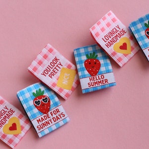 May include: Six colorful fabric labels with gingham patterns and embroidered strawberries. The labels feature phrases like "Lovingly Handmade", "You Look Pretty", "Made for Sunny Days", and "Hello Summer".
