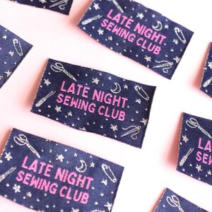 May include: Navy blue fabric labels with the text "LATE NIGHT SEWING CLUB" in pink. The labels are decorated with silver-coloured scissors, needles, stars, and crescent moons. The labels are scattered on a pink surface.