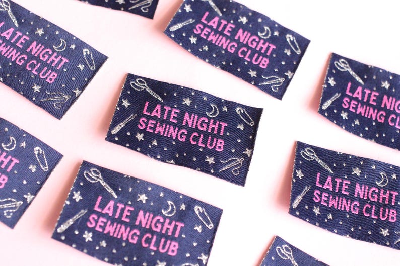 May include: Navy blue fabric labels with the text "LATE NIGHT SEWING CLUB" in pink. The labels are decorated with silver-coloured scissors, needles, stars, and crescent moons. The labels are scattered on a pink surface.