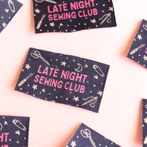 May include: Navy blue fabric labels with the text "LATE NIGHT. SEWING CLUB" in pink. The labels are decorated with white illustrations of scissors, needles, thread, stars, and moons. The labels are scattered on a pink surface.