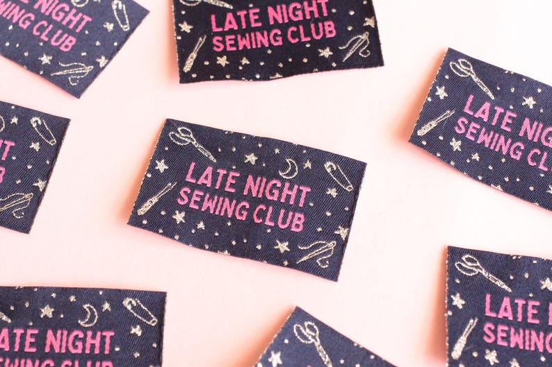 May include: Navy blue fabric labels with the text "LATE NIGHT. SEWING CLUB" in pink. The labels are decorated with white illustrations of scissors, needles, thread, stars, and moons. The labels are scattered on a pink surface.