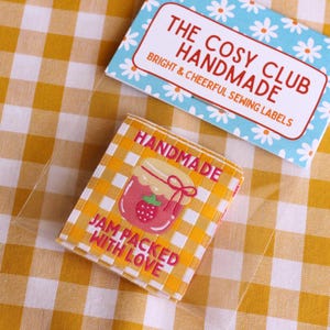 May include: Two labels on a yellow and white checkered surface. One label is light blue with white daisies and reads "THE COSY CLUB HANDMADE BRIGHT & CHEERFUL SEWING LABELS". The other label is yellow and white checkered with a strawberry jam jar graphic and reads "HANDMADE JAM PACKED WITH LOVE".