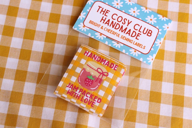May include: Two labels on a yellow and white checkered surface. One label is light blue with white daisies and reads "THE COSY CLUB HANDMADE BRIGHT & CHEERFUL SEWING LABELS". The other label is yellow and white checkered with a strawberry jam jar graphic and reads "HANDMADE JAM PACKED WITH LOVE".