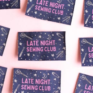 May include: Several navy blue fabric patches, each embroidered with the text "LATE NIGHT. SEWING CLUB" in pink. The patches feature embroidered stars, moons, scissors, needles, and pins. They are arranged on a pale pink background.