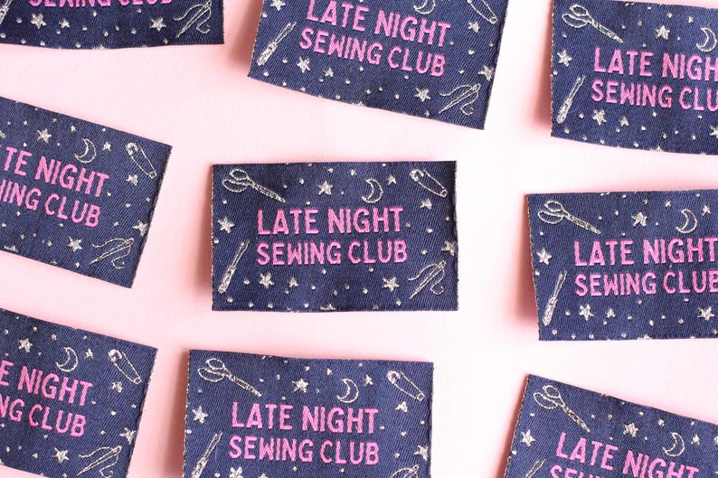 May include: Several navy blue fabric patches, each embroidered with the text "LATE NIGHT. SEWING CLUB" in pink. The patches feature embroidered stars, moons, scissors, needles, and pins. They are arranged on a pale pink background.