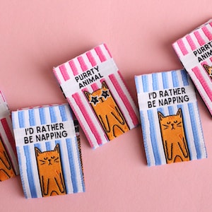 May include: Four colorful iron-on patches with cat illustrations and text. The patches feature a pink and white striped background with the text "Purrty Animal" and a cat wearing sunglasses, a blue and white striped background with the text "I'd rather be napping" and a cat with closed eyes, and a pink and white striped background with the text "Purrty Animal" and a cat wearing sunglasses.