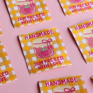 May include: Close-up of several rectangular labels with a yellow and white checkered pattern. Each label features a pink jar of jam with a strawberry and the text "HANDMADE" and "JAM PACKED WITH LOVE" in pink.
