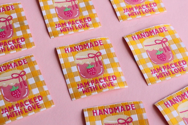 May include: Close-up of several rectangular labels with a yellow and white checkered pattern. Each label features a pink jar of jam with a strawberry and the text "HANDMADE" and "JAM PACKED WITH LOVE" in pink.
