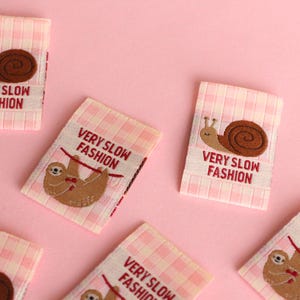Peut inclure: Cinq &eacute;tiquettes en tissu vichy rose et blanc avec des images brod&eacute;es d'un escargot et d'un paresseux. Les &eacute;tiquettes portent le texte "VERY SLOW FASHION".