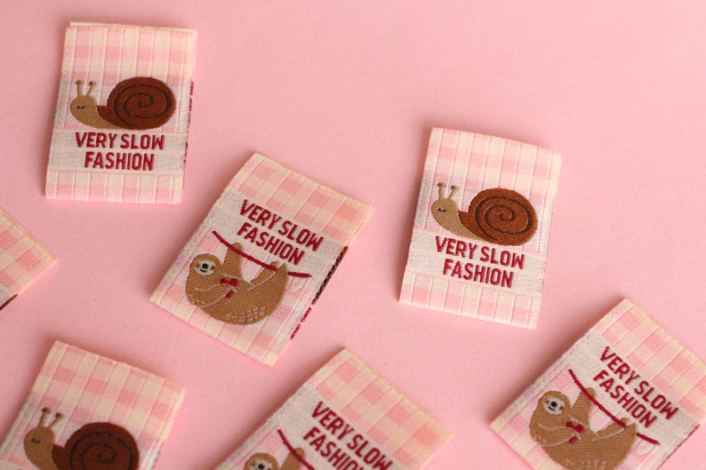 Peut inclure: Cinq &eacute;tiquettes en tissu vichy rose et blanc avec des images brod&eacute;es d'un escargot et d'un paresseux. Les &eacute;tiquettes portent le texte "VERY SLOW FASHION".