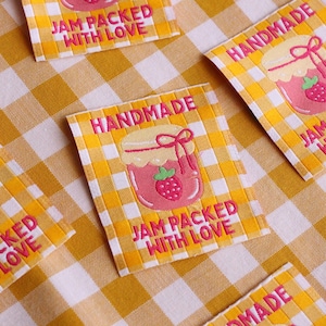 May include: Several fabric labels with a yellow and white checkered background. Each label features a pink jar of jam with a strawberry and the words "HANDMADE" and "JAM PACKED WITH LOVE" in pink.