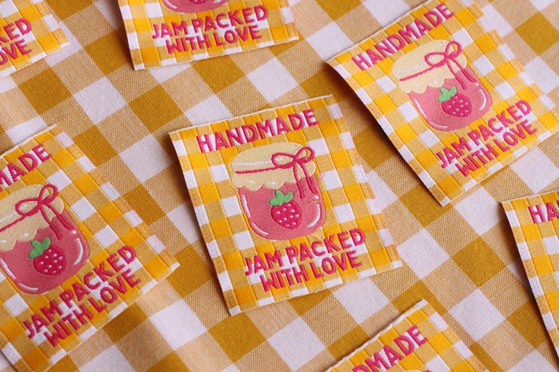May include: Several fabric labels with a yellow and white checkered background. Each label features a pink jar of jam with a strawberry and the words "HANDMADE" and "JAM PACKED WITH LOVE" in pink.