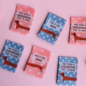 May include: A collection of small, rectangular fabric patches. Some are pink with white hearts, others are blue with white polka dots. Each features a brown dachshund illustration and text that reads "THIS IS THE BACK" or "THIS TOOK A LOOOOOONG TIME TO MAKE."