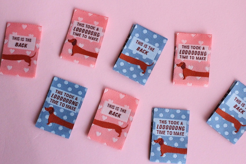 May include: A collection of small, rectangular fabric patches. Some are pink with white hearts, others are blue with white polka dots. Each features a brown dachshund illustration and text that reads "THIS IS THE BACK" or "THIS TOOK A LOOOOOONG TIME TO MAKE."