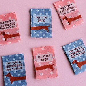 May include: Small rectangular fabric labels in pink and blue with white polka dots and hearts. Each label features a dachshund illustration and text that reads "THIS TOOK A LOOOOOONG TIME TO MAKE" or "THIS IS THE BACK".
