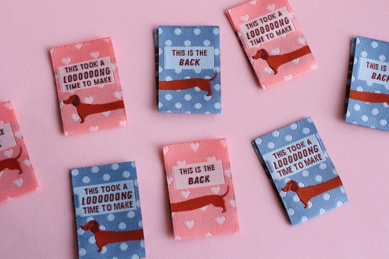 May include: Small rectangular fabric labels in pink and blue with white polka dots and hearts. Each label features a dachshund illustration and text that reads "THIS TOOK A LOOOOOONG TIME TO MAKE" or "THIS IS THE BACK".