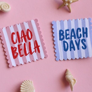 Op de afbeelding: Twee vierkante kaarten met geschulpte randen, één met roze en witte strepen en de woorden "CIAO BELLA" in rood, de andere met blauw-witte strepen en de woorden "BEACH DAYS" in blauw. Schelpen en zeesterren op een roze achtergrond.