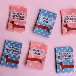 May include: Rectangular fabric labels with dachshund illustrations and text. Some labels are pink with white hearts, others are blue with white polka dots. Text includes "THIS IS THE BACK" and "THIS TOOK A L000000NG TIME TO MAKE."