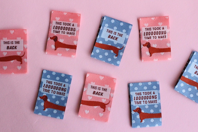May include: Rectangular fabric labels with dachshund illustrations and text. Some labels are pink with white hearts, others are blue with white polka dots. Text includes "THIS IS THE BACK" and "THIS TOOK A L000000NG TIME TO MAKE."