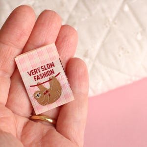Peut inclure: Une &eacute;tiquette en tissu vichy rose et blanc avec un paresseux brun suspendu &agrave; une ficelle rouge. L'&eacute;tiquette indique "Very Slow Fashion".