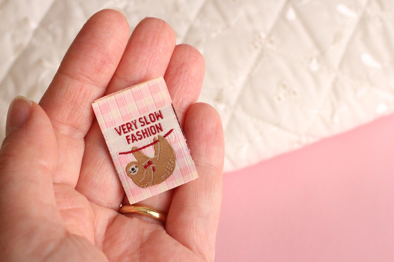 Peut inclure: Une &eacute;tiquette en tissu vichy rose et blanc avec un paresseux brun suspendu &agrave; une ficelle rouge. L'&eacute;tiquette indique "Very Slow Fashion".