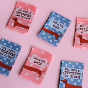 May include: Fabric labels with dachshund illustrations and text. Some labels are pink with white hearts, others are blue with white polka dots. Text includes "This is the back" and "This took a loooooong time to make."