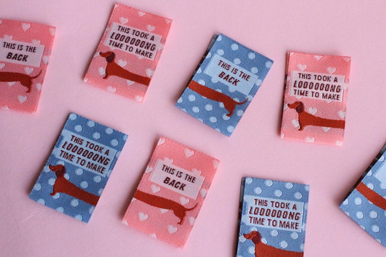 May include: Fabric labels with dachshund illustrations and text. Some labels are pink with white hearts, others are blue with white polka dots. Text includes "This is the back" and "This took a loooooong time to make."
