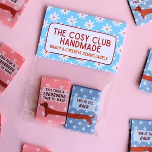 May include: Sewing labels with dachshund illustrations. The labels are pink and blue with white polka dots and text. The labels read "This took a L0000000NG time to make" and "This is the back". The packaging reads "The Cosy Club Handmade".