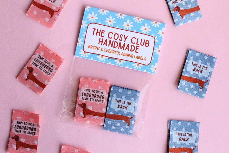 May include: Sewing labels with dachshund illustrations. The labels are pink and blue with white polka dots and text. The labels read "This took a L0000000NG time to make" and "This is the back". The packaging reads "The Cosy Club Handmade".