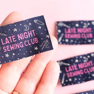 May include: A navy blue fabric label with the text "LATE NIGHT SEWING CLUB" in pink. The label is decorated with silver-coloured sewing-related icons, including scissors, needles, and stars. The label is held in a hand.