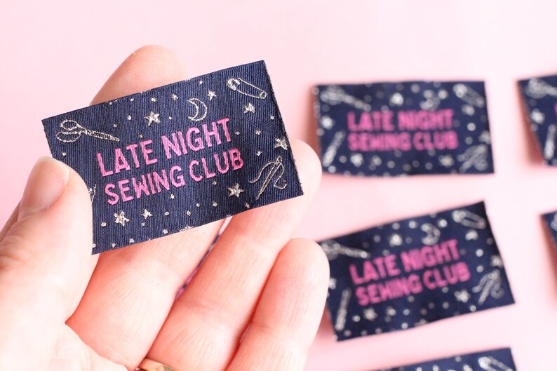 May include: A navy blue fabric label with the text "LATE NIGHT SEWING CLUB" in pink. The label is decorated with silver-coloured sewing-related icons, including scissors, needles, and stars. The label is held in a hand.