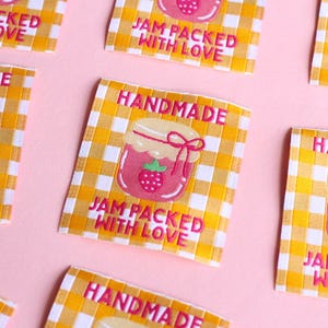 May include: Several fabric labels with a yellow and white checkered pattern and the words "HANDMADE" and "JAM PACKED WITH LOVE" in pink. Each label features a pink jam jar with a strawberry.