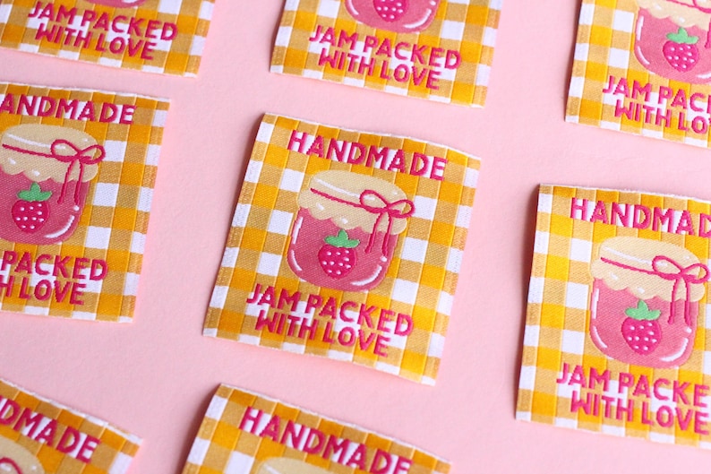 May include: Several fabric labels with a yellow and white checkered pattern and the words "HANDMADE" and "JAM PACKED WITH LOVE" in pink. Each label features a pink jam jar with a strawberry.