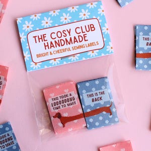 May include: Sewing labels with dachshund illustrations. The labels are in pink and blue with white polka dots and hearts. Text on the labels reads "This is the back" and "This took a loooong time to make". The packaging reads "The Cosy Club Handmade".
