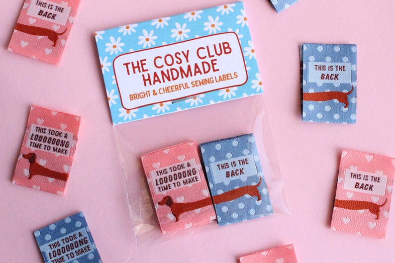 May include: Sewing labels with dachshund illustrations. The labels are in pink and blue with white polka dots and hearts. Text on the labels reads "This is the back" and "This took a loooong time to make". The packaging reads "The Cosy Club Handmade".