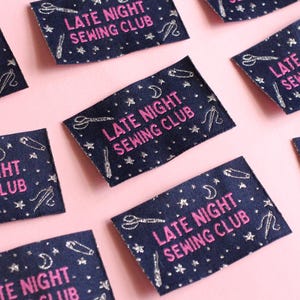 May include: Navy blue fabric labels with the text "LATE NIGHT SEWING CLUB" in pink. The labels are decorated with silver-coloured scissors, pins, moons, and stars. The labels are arranged on a pink background.