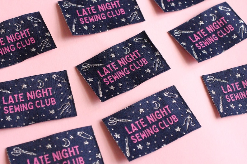 May include: Navy blue fabric labels with the text "LATE NIGHT SEWING CLUB" in pink. The labels are decorated with silver-coloured scissors, pins, moons, and stars. The labels are arranged on a pink background.