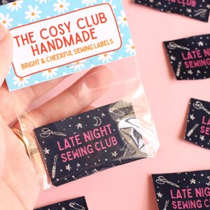 May include: A hand holds a clear bag containing navy blue fabric labels. Each label features white sewing tool illustrations and the pink text "LATE NIGHT SEWING CLUB". The bag's top has a blue and white floral pattern, with the text "THE COSY CLUB HANDMADE".