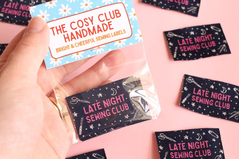 May include: A hand holds a clear bag containing navy blue fabric labels. Each label features white sewing tool illustrations and the pink text "LATE NIGHT SEWING CLUB". The bag's top has a blue and white floral pattern, with the text "THE COSY CLUB HANDMADE".