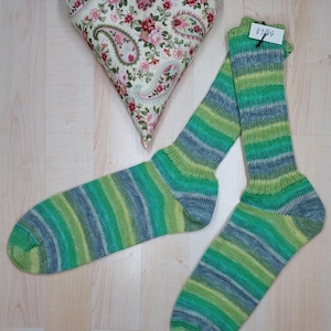 Könnte beinhalten: Ein Paar grün, blau und grau gestreifte Socken mit einem herzförmigen Kissen. Das Kissen hat ein florales Paisley-Muster. Die Socken sind handgefertigt und scheinen gestrickt zu sein. Auf einer hellen Holzoberfläche.