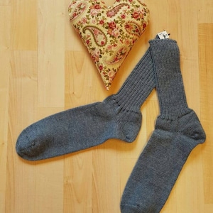 Könnte beinhalten: Ein Paar graue Strickstrümpfe mit Etikett, neben einem herzförmigen Kissen mit floralem Muster. Die Socken liegen auf einer hellbraunen Holzoberfläche.