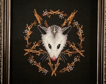 May include: A framed cross-stitch embroidery featuring a white opossum with a pentagram on its forehead, surrounded by a golden wreath of thorns. The background is black.
