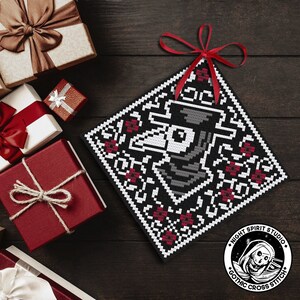 May include: Black and white cross stitch pattern of a plague doctor's mask with red accents. The pattern is framed in a black square with a white border. The image includes the text "Night Spirit Studio Gothic Cross Stitch".