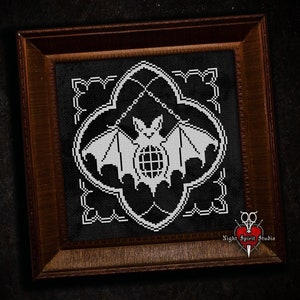 May include: Black and white cross stitch pattern of a bat in a decorative frame. The bat is in the center of the frame and has a stylized design. The frame is made of wood and has a dark brown finish. The text "Night Spirit Studio" is in the bottom right corner of the image.