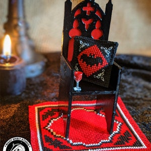 May include: A miniature black gothic chair with a red cushion and a bat-shaped pillow. A small glass of red liquid sits on a shelf on the chair. The chair is on a red and black rug with a white border. The rug has a fringe border. The image includes the text "Night Spirit Studio Gothic Cross Stitch".