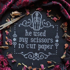 May include: A black cross-stitch with the text "he used my scissors to cut paper" in white. The design includes a skeleton and decorative flourishes. Silver scissors and dried rose petals surround the cross-stitch, set against a dark red and black fabric.