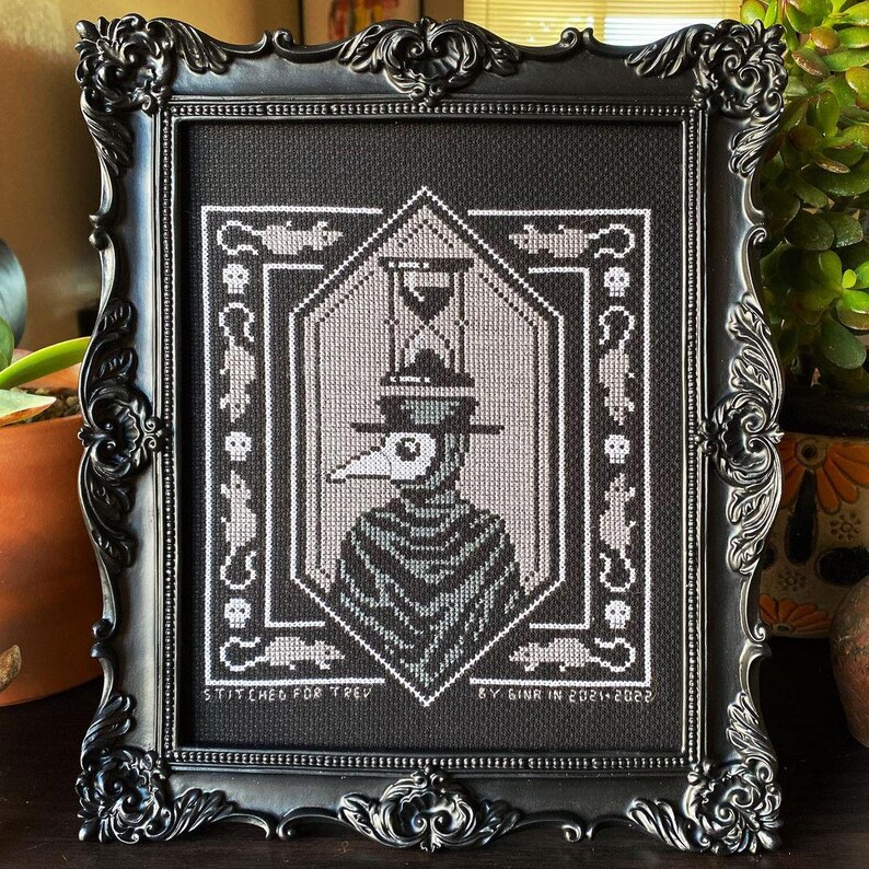 May include: Black and white cross-stitch embroidery in a black frame. The design features a figure wearing a plague doctor mask and hat, with an hourglass above. The text "Stitched for Trey by Ginr in 2021-2022" is at the bottom.