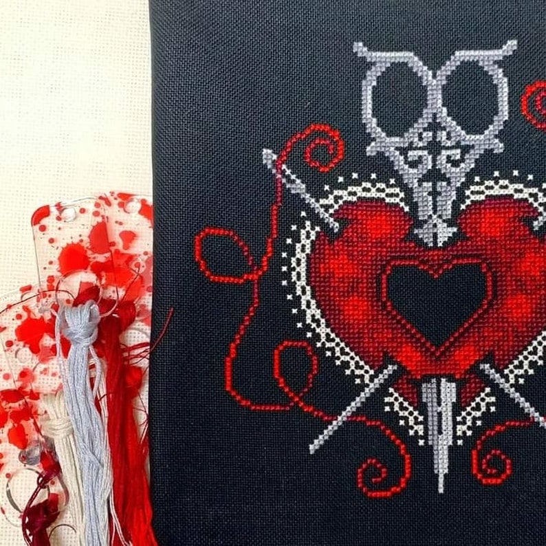 Puede incluir: Patr&oacute;n de punto de cruz de un coraz&oacute;n rojo atravesado por una daga y dos agujas, rodeado de un dise&ntilde;o de encaje blanco sobre un fondo negro.