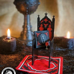 K&ouml;nnte beinhalten: Ein Miniatur-Thron aus schwarzem Holz mit einem roten Samtkissen und einem kleinen Glas mit roter Fl&uuml;ssigkeit. Der Thron steht auf einem roten und schwarzen Teppich mit Muster. Im Hintergrund sind ein schwarzer Kelch und zwei brennende Kerzen zu sehen.
