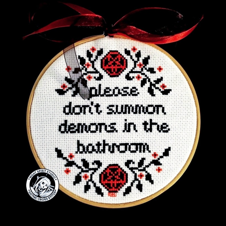 May include: A white embroidery hoop with black and red thread. The embroidery reads "please don't summon demons in the bathroom". The hoop is decorated with red and black pentagrams and floral designs. The bottom of the hoop has a black and white logo that reads "Night Spirit Studio Gothic Cross Stitch".